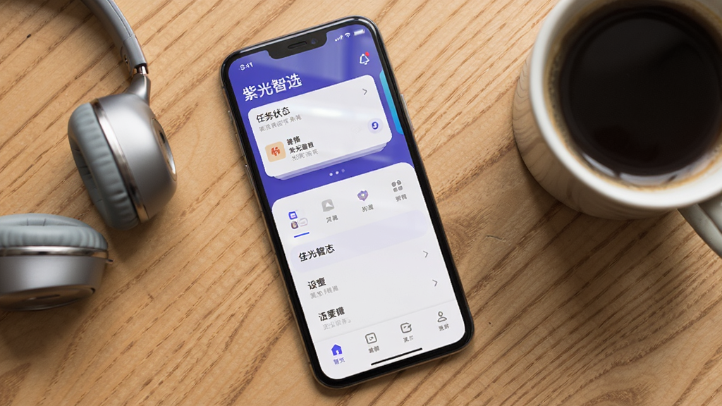 画面中是一部智能手机,屏幕上显示「紫光智选」移动端界面,界面顶部有任务状态卡片,下方是简洁的菜单栏,手机旁边放着一副耳机和一杯咖啡,布局采用俯视角度排版,风格为清新生活风,细节包括手机屏幕上的微光反射、桌面木纹纹理,画质清晰干净,无多余元素