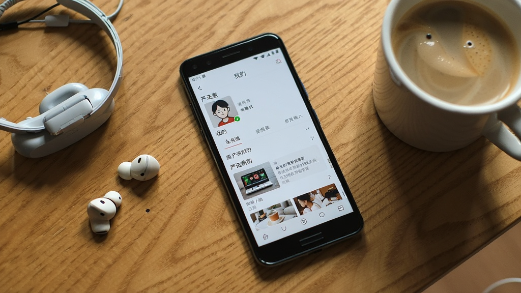 一部安卓手机放置在桌面上,屏幕显示严选惠APP的“我的”页面,旁边放着耳机和一杯咖啡,背景为木质桌面,风格生活化,画质清晰自然,无多余元素