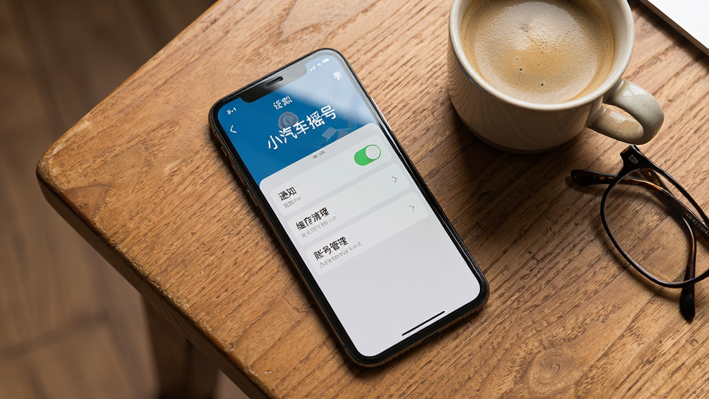 一部手机放置在木质桌面上,屏幕亮起显示“小汽车摇号”应用的设置页面,页面上有通知开关、缓存清理、账号管理等选项,手机旁边有一杯咖啡和一副眼镜,营造出日常办公或居家使用的场景。布局为俯视角度拍摄风格,色彩温暖自然,细节上手机屏幕反光清晰,桌面纹理可见,画质高清,画面干净无杂乱物品。