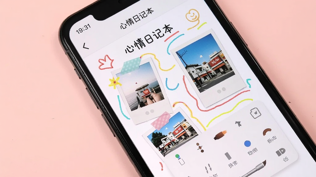 一部手机屏幕显示「心情日记本」的编辑界面,画面中有手绘涂鸦的彩色线条和插入的照片缩略图,背景为浅粉色,风格活泼可爱,排版自由,细节上画笔工具条清晰可见,画质清晰,干净,无多余元素