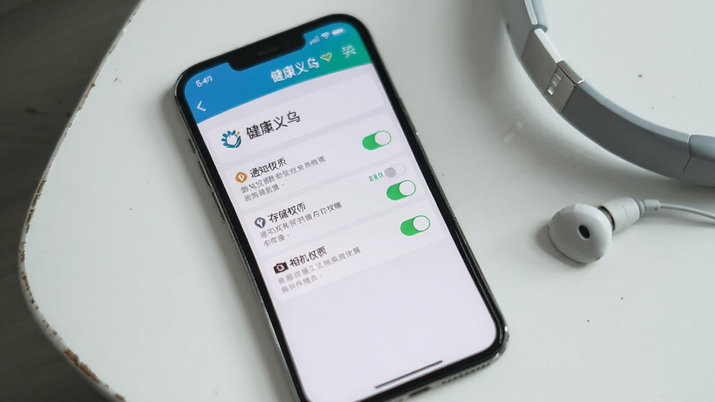 一张展示手机屏幕上“健康义乌”软件权限设置界面的图片,界面中包含通知权限、存储权限与相机权限的开关选项,手机放置于白色桌面,旁边有一副耳机,背景简洁,风格写实,细节清晰,画质高清,无多余元素