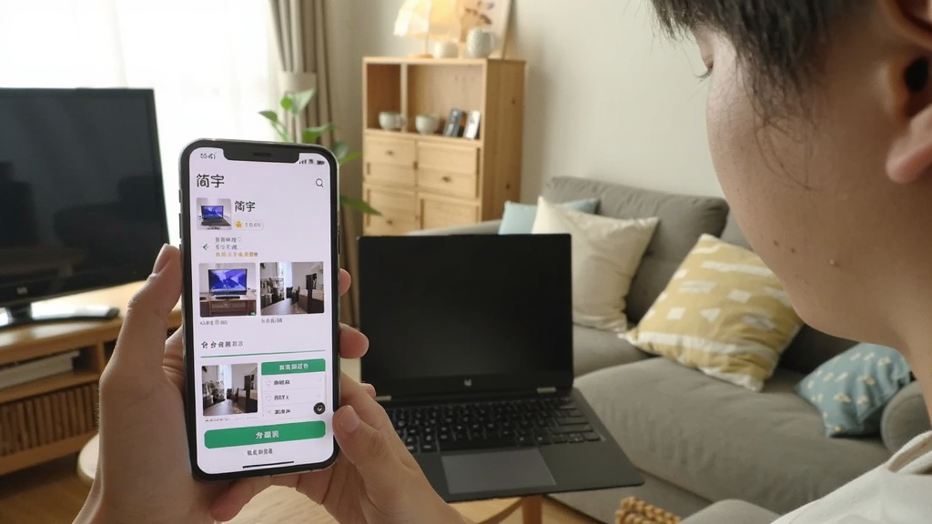 一个年轻人在家中客厅用手机拍摄闲置的笔记本电脑,手机屏幕显示“简宇”发布商品页面,页面中有照片预览和分类选择按钮,背景是温馨的家居环境,画面明亮自然,风格生活化,细节丰富,画质清晰,干净无多余元素