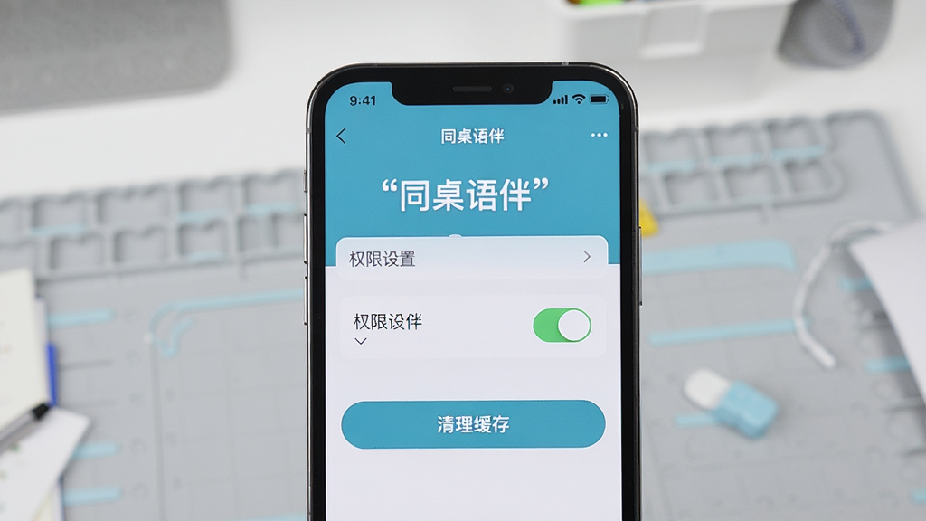 主体是一个手机屏幕特写,显示同桌语伴的设置页面,页面中有权限开关和清理缓存按钮;布局为手机竖屏居中,背景为浅灰色工作台;排版清晰,风格为写实产品摄影;细节上屏幕内需有引号“同桌语伴”和引号“权限设置”字样;画质高清,画面干净,光线均匀。