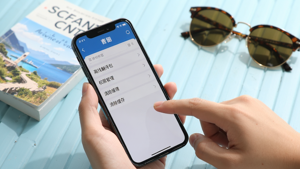 一部手机展示出国翻译官的设置页面,屏幕内可见“离线翻译包”“权限管理”“清除缓存”等选项,手机旁边有一只手正在点击屏幕。背景为浅蓝色桌面,放置着一本旅行指南和一副墨镜,整体风格清新明亮,构图自然,画质清晰,无多余元素。