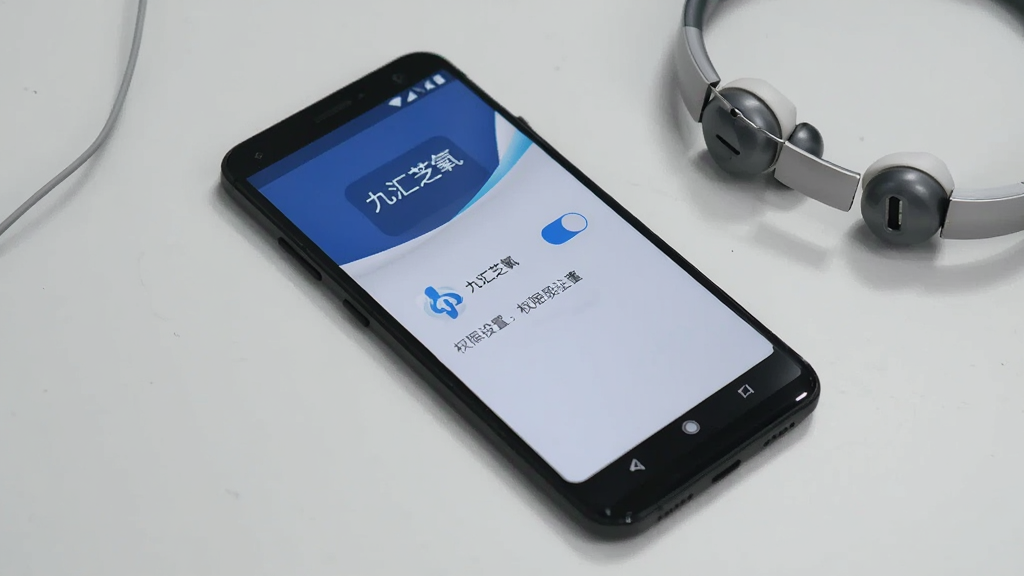 画面主体为一部安卓手机,屏幕上显示九汇芝氧的权限设置引导页面,页面中有两个开关按钮与说明文字,手机旁边放着一副耳机。布局为斜侧视角,背景为浅灰色桌面。风格为数码产品展示风,细节上手机屏幕有轻微反光,应用名称“九汇芝氧”显示在页面顶部。画质清晰,干净,无多余元素。