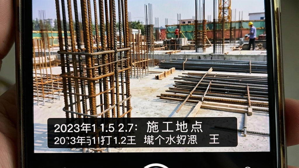水印相机打卡王App界面特写,画面主体为手机屏幕中一张刚拍好的工地照片,照片底部叠加半透明黑色水印条,清晰显示日期时间与施工地点文字,布局为照片占屏幕大部分、水印条横贯底部,风格为真实手机截图风,细节包括水印文字的清晰边缘与照片中的钢筋建材,画质高清,干净无多余元素