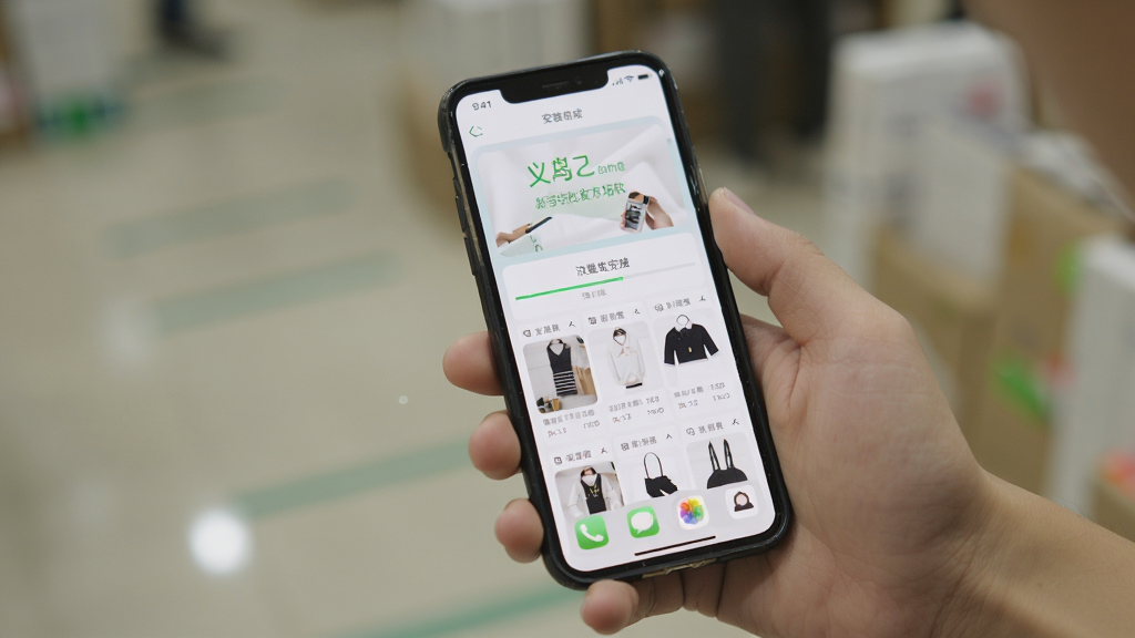 义乌特价批发市场软件新手安装使用场景图,主体为手持手机展示安装完成界面与商品浏览页面,布局为人物手部特写加手机屏幕内容,排版简洁聚焦,风格为生活化实拍感,细节包含手机桌面图标、安装进度条、商品列表界面,画质清晰干净无多余背景元素
