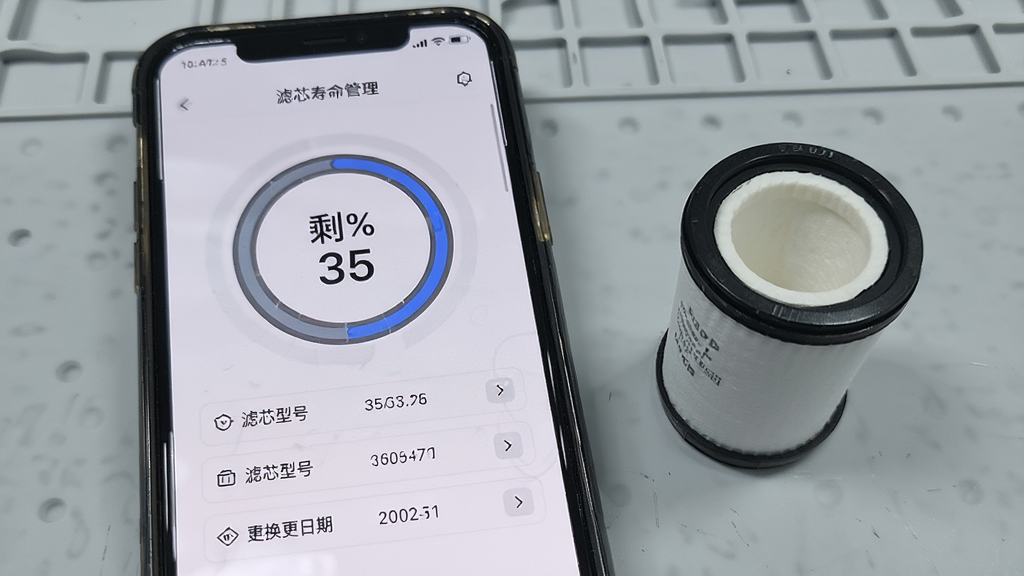 画面展示造梦者软件内滤芯寿命管理页面,手机屏幕中央有一个环形进度条显示剩余百分之三十五,下方列出滤芯型号与建议更换日期,旁边放置一枚真实的圆筒形滤芯实物作为参照,背景为浅灰色工作台面,整体风格偏向科技感与实用感结合,光线均匀,画质清晰,画面简洁