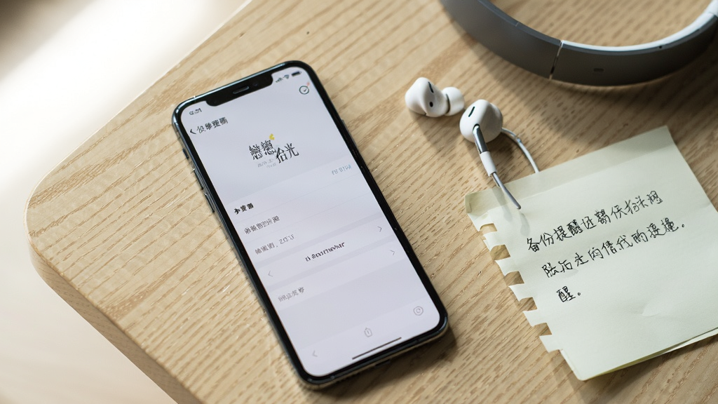 一部手机平放在桌面上,屏幕显示“拾光”的设置页面,旁边放着一副耳机和一张写有备份提醒的便签纸,桌面为浅色木纹,光线从左上角照入,风格生活化,画质清晰干净,构图简洁
