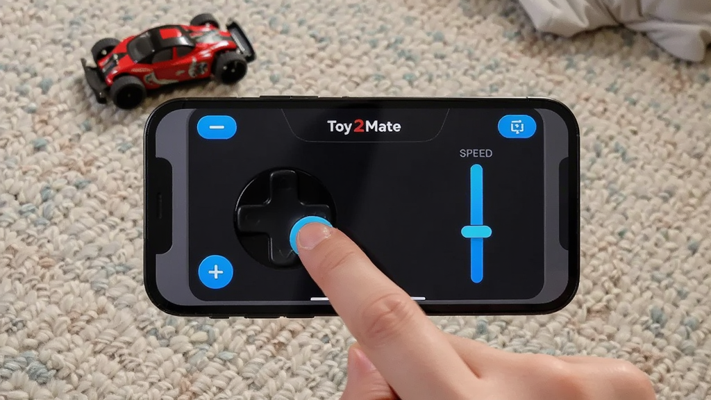 画面主体是一部手机横屏显示“Toy2Mate”遥控面板,面板上有虚拟摇杆和速度滑块,布局为手机居中、背景是客厅地毯与玩具小车,排版突出横屏操控界面,风格为生活场景实拍风,细节包含手指放在摇杆上的动作、屏幕中的蓝色高亮控件、地毯纹理清晰,画质高清、干净、无多余元素