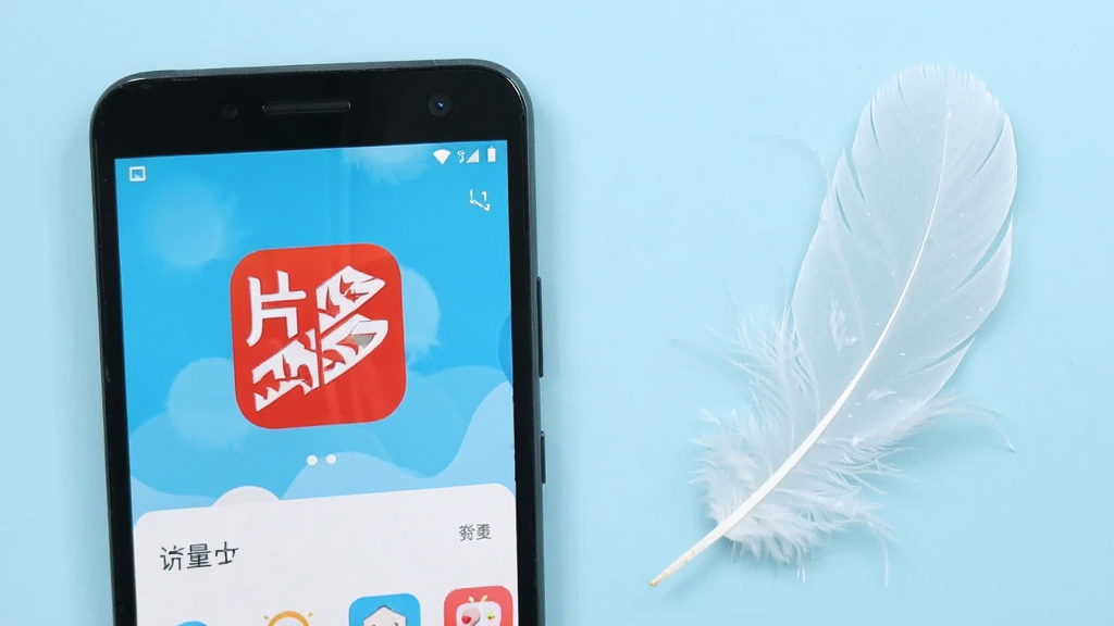 画面展示一部中低端安卓手机与一片轻盈的羽毛并排摆放,手机屏幕显示片多多应用图标和流畅运行的界面,羽毛象征轻量,背景为浅蓝色纯色,风格简约清爽,布局左重右轻形成对比,画质干净利落无多余装饰