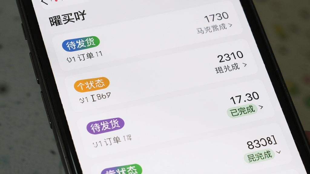 画面主体是手机屏幕特写,显示“曜买吖”的订单列表页面,多条订单卡片纵向排列,每张卡片上有状态标签和金额信息;布局为手机屏幕充满画面,突出界面内容;排版上订单卡片间距均匀,状态标签用不同颜色区分;风格为扁平化UI展示风格;细节上金额数字清晰,状态文字“待发货”“已完成”可辨认;画质锐利干净,无多余装饰元素。
