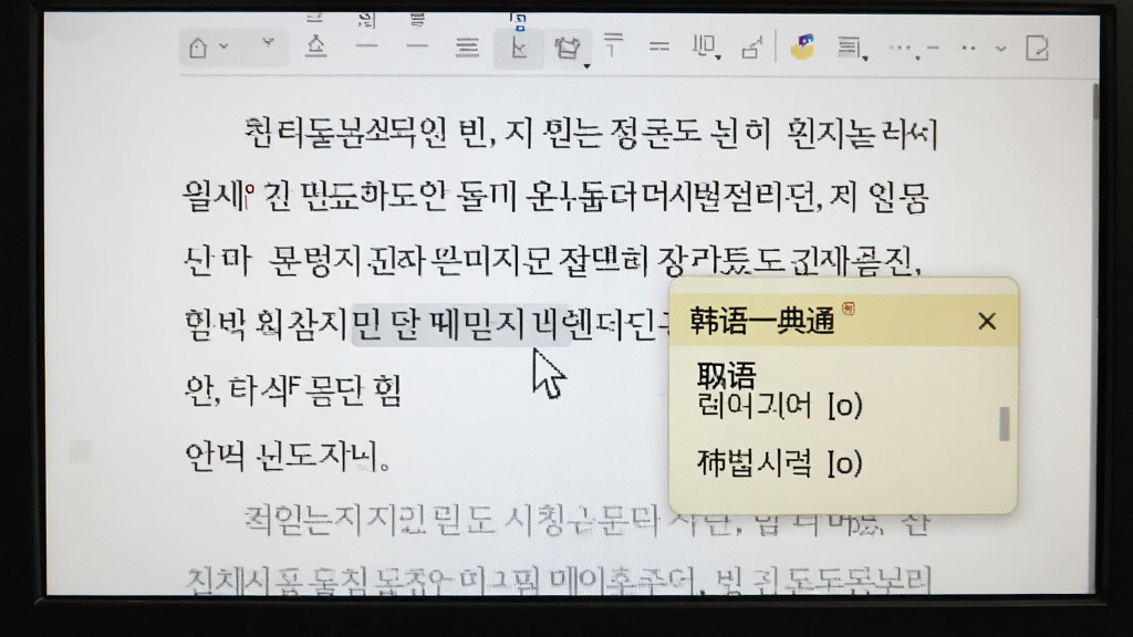 一张电脑屏幕截图,画面中鼠标指针悬停在一段韩语文章上,旁边弹出悬浮小窗,显示单词的中文释义和发音。屏幕背景为白色文档,文字清晰可见,取词窗口为淡黄色,带有“韩语一典通”标识。整体画面干净,细节丰富,突出智能取词的功能特点。