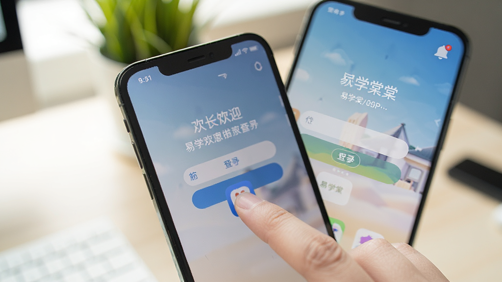 展示易学棠APP在手机桌面上运行的实拍场景,画面中有手指正点击应用图标,屏幕上显示登录后的欢迎页面,右上角有消息铃铛图标,整体光线明亮,背景为书桌与绿植虚化效果,强调真实使用状态,画质干净清晰。
