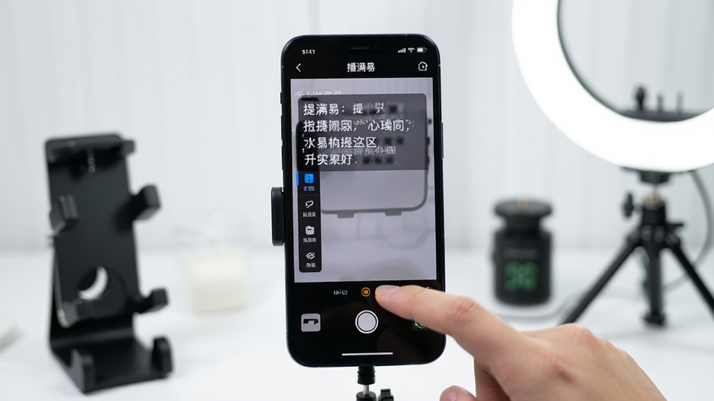 主体:一部手机正在进行“播满易”软件的实际操作演示,屏幕显示提词与拍摄同屏界面。布局:手机位于画面中央,周围散落着手机支架、补光灯等配件。排版:屏幕上可见清晰的提词文字与底部拍摄按钮,左侧有功能菜单栏。风格:实拍操作场景,浅景深效果,背景干净整洁。细节:手指点击屏幕的瞬间被捕捉,画面生动真实。画质:高清,干净,无多余元素,直观体现软件操作流畅性。