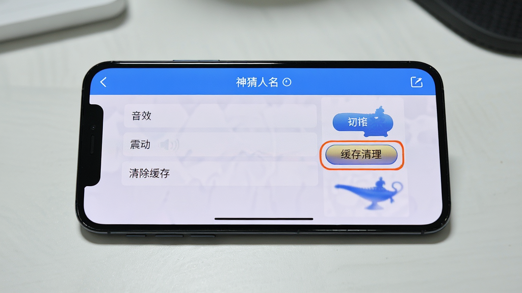 手机横屏放置在桌面上,屏幕上显示神灯猜人名的设置界面,有“音效”、“震动”、“清除缓存”等选项。主体:横屏手机与设置界面。布局:居中构图。排版:设置列表清晰。风格:实拍风格。细节:高亮显示“缓存清理”按钮,画质高清,桌面整洁。
