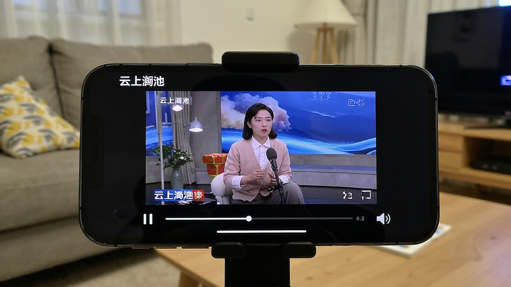 一张展示手机视频播放界面的图片,画面中手机横屏显示“云上渑池”的视频播放页面,屏幕中有本地电视节目的画面,下方有播放进度条和音量图标。背景是客厅沙发环境,手机支架固定,氛围轻松休闲,画质清晰,突出视听娱乐功能。