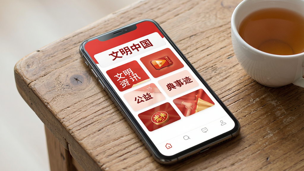 一张手机屏幕展示文明中国应用主界面,界面顶部有文明中国四个字标题,下方排列着文明资讯、公益广告、典型事迹等卡片式栏目入口,整体配色以红色和白色为主,背景干净简洁,手机放置在一张木质桌面上,旁边有一杯茶,风格写实,细节清晰,画质高清,干净,无多余元素
