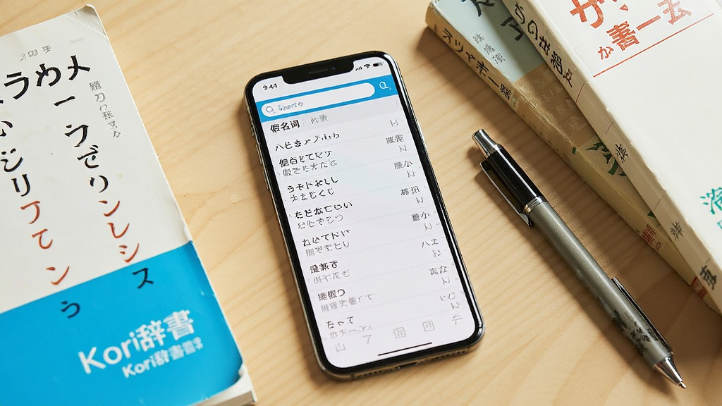 画面中央展示一部智能手机,手机屏幕显示日语词典查询界面,界面顶部有搜索框,中间排列日语假名词条与中文释义,手机旁边放置几本日语学习书籍和一支笔,背景为浅色木质桌面,整体布局简洁干净,风格为写实产品展示风,细节包括屏幕反光、书本纹理、笔杆阴影,画质清晰,无多余元素,画面中文字标注“Kori辞书”
