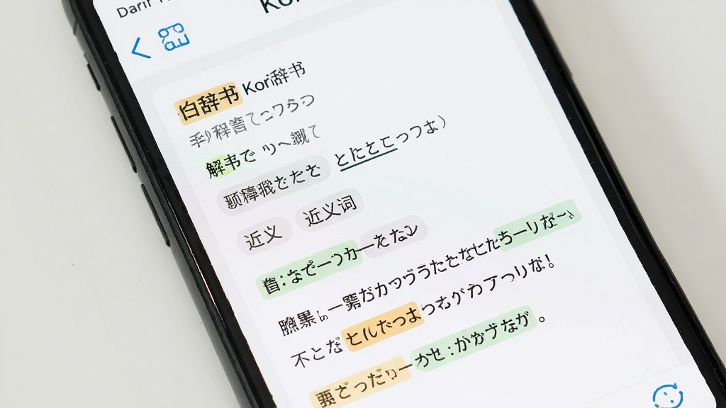 画面展示手机屏幕显示日语词条详情页,页面包含单词、释义、多条例句以及近义词标签,例句中关键词高亮显示,背景为白色卡片式布局,风格为现代词典应用界面展示,细节包括高亮色块、标签圆角、字体层级,画质清晰,无多余元素,画面中文字标注“Kori辞书”