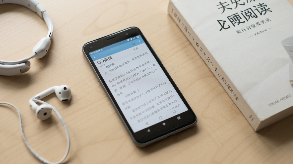一部安卓手机放在桌面上,屏幕显示“QQ阅读”阅读界面,旁边有耳机和一本纸质书,背景是简洁的浅色桌面,光线自然,风格生活化,细节清晰,画质干净,无多余元素