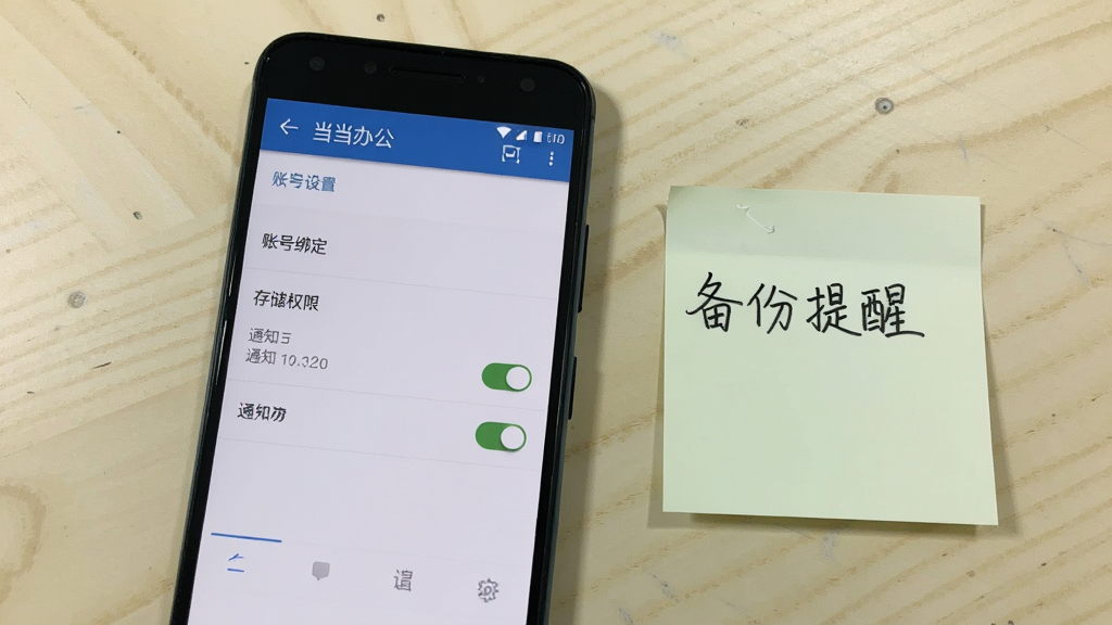 一部安卓手机显示当当办公的设置页面,页面中包含账号绑定、存储权限、通知开关等选项,手机旁边放着一张手写的便签纸,上面写着备份提醒,背景是浅色桌面,风格为实用教程风,画面清晰,画质干净,无多余元素
