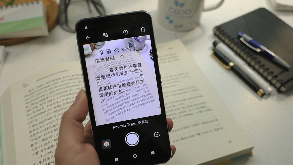 一张展示安卓翻译官拍照翻译功能的图片,画面中一部安卓手机正在拍摄一本外文书籍,手机屏幕上显示着翻译后的中文文字,背景是书桌和文具,风格清新,细节丰富,画质干净,无多余元素