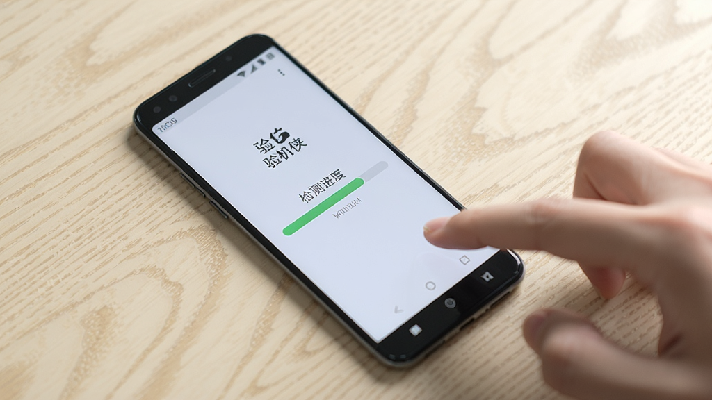一部安卓手机放置在桌面上,屏幕显示验机侠的检测进度条,手机旁边有一只手正在点击屏幕,背景为浅色木纹桌面,画面风格写实清新,光线柔和,画质干净,画面中可见“验机侠”字样,无多余元素