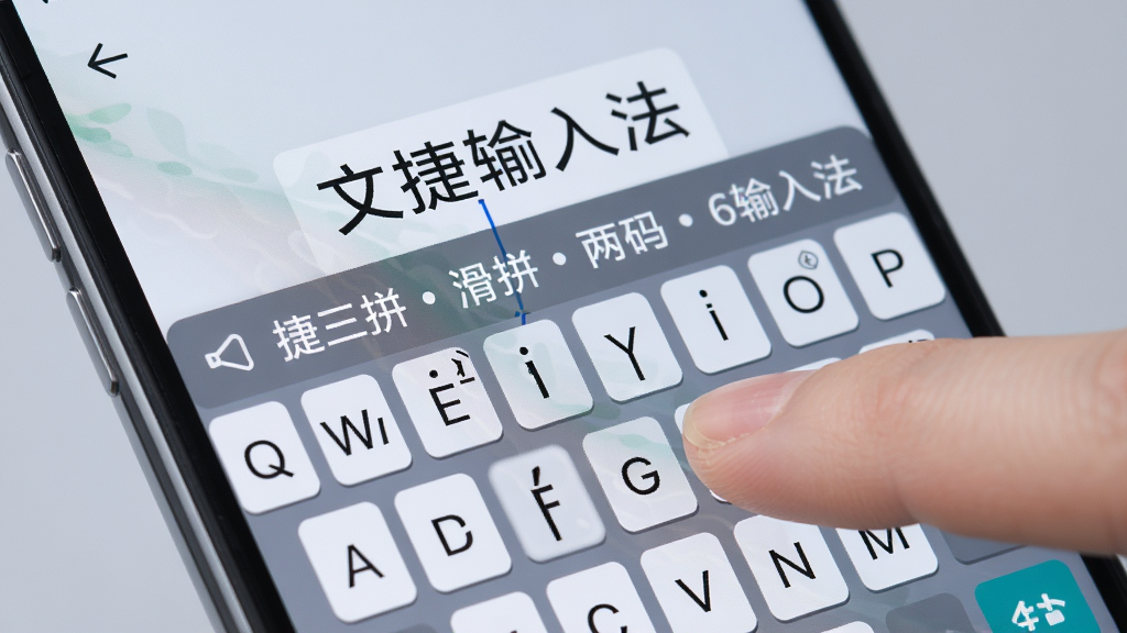 一部手机屏幕特写,屏幕上显示文捷三拼滑拼两码6笔输入法的键盘区域,有手指正在滑动输入,键盘上方浮现出候选词“文捷输入法”,背景为柔和的浅灰色,画面风格写实,画质清晰,细节丰富,无多余元素