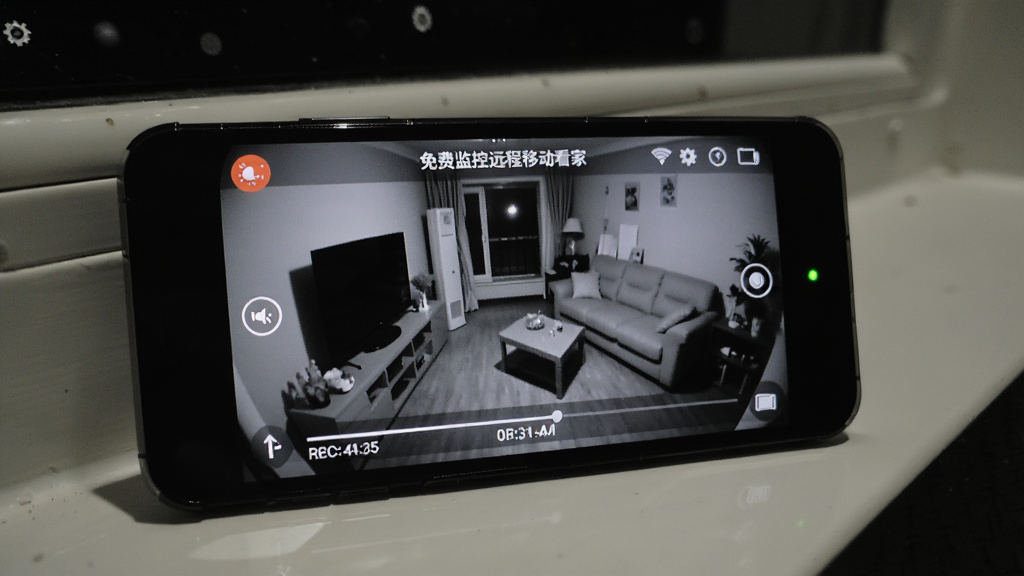 一部手机放置在窗台边作为监控端,屏幕显示“免费监控远程移动看家”的夜视画面,画面中呈现灰白色的夜间客厅景象,风格为暗光摄影,布局为手机特写,细节包括屏幕上有红外夜视图标、录像时间轴、以及微弱的绿色指示灯,画质清晰,干净无多余元素