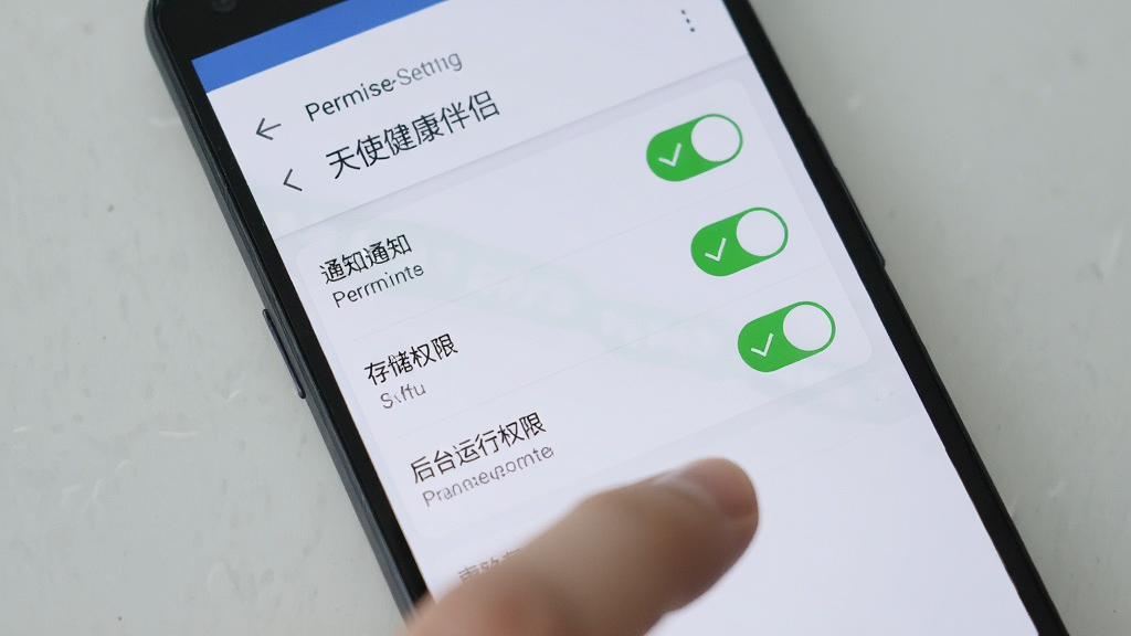 一部安卓手机屏幕显示“天使健康伴侣”的权限设置页面,页面中通知权限、存储权限、后台运行权限三个开关均处于开启状态,开关旁边有绿色对勾标记;手机下方有一只手正在点击屏幕;背景为浅灰色桌面;风格写实,细节清晰;画质干净,无多余元素