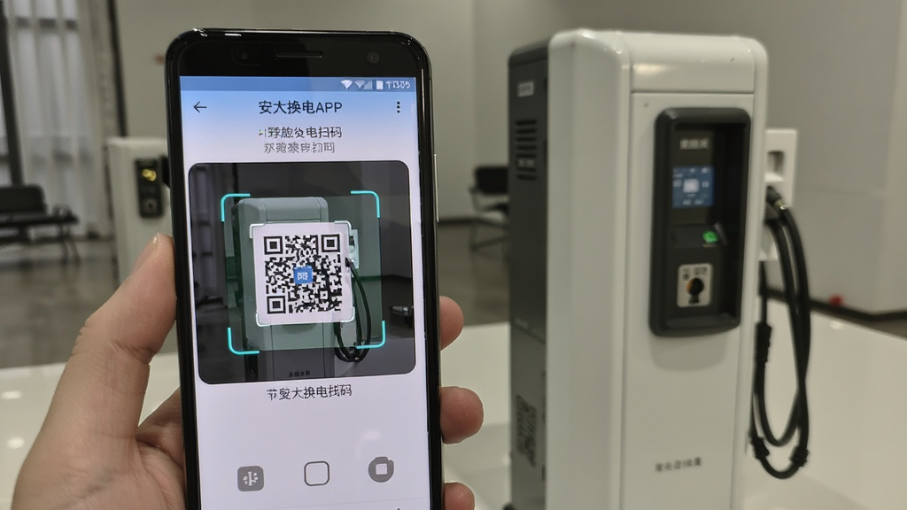 一张展示安达换电APP在安卓手机上操作界面的实拍图,画面中手机屏幕显示换电扫码页面,摄像头对准换电站二维码,屏幕上有扫描框和提示文字。手机旁边放置一个换电站设备模型,背景为室内场景,整体风格真实自然,画质清晰,适合用于软件使用教程配图。