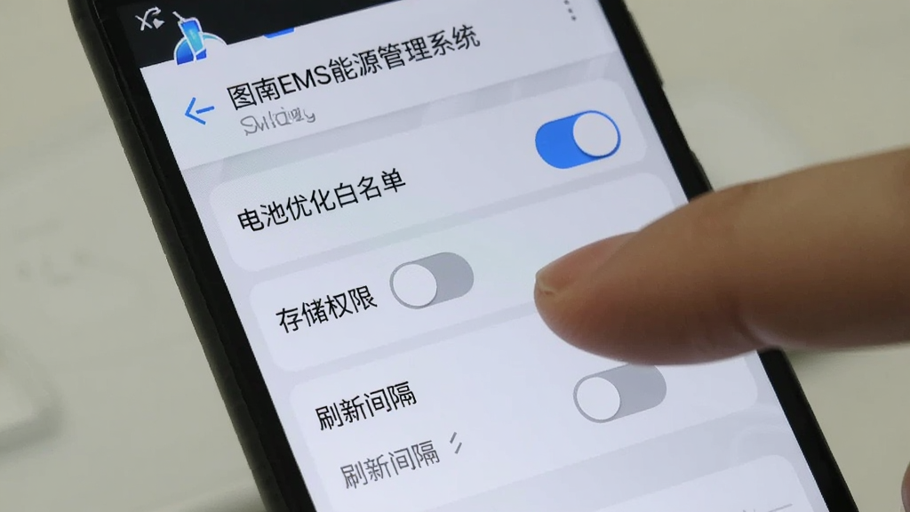 一部安卓手机屏幕显示“图南EMS能源管理系统”的设置页面,页面中有电池优化白名单开关、存储权限开关与刷新间隔选项,背景为浅色主题,细节包含手指点击屏幕的动作与设置图标,画质清晰,干净,无多余元素