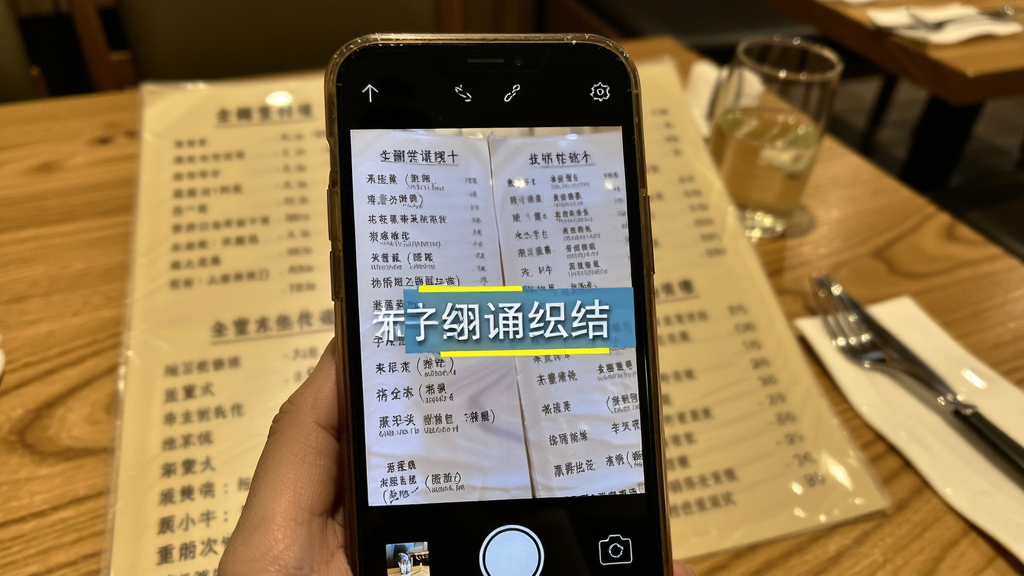 主体为AI翻译机拍照翻译功能展示,布局为手机摄像头对准外文菜单,屏幕上叠加中文翻译浮层,排版主体突出手机与菜单,风格为写实生活场景,细节包括餐厅桌面、菜单文字、翻译高亮框,画质清晰干净无多余元素