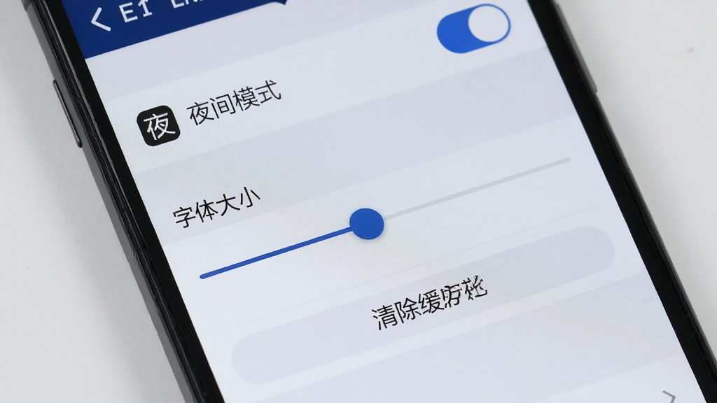 一张展示墨绘软件设置菜单界面的手机特写图,画面中手机屏幕显示着设置列表,包含夜间模式开关、字体大小滑块、清除缓存按钮等选项,界面布局清晰,图标简约,背景为浅灰色,光线均匀,画质清晰,无多余元素