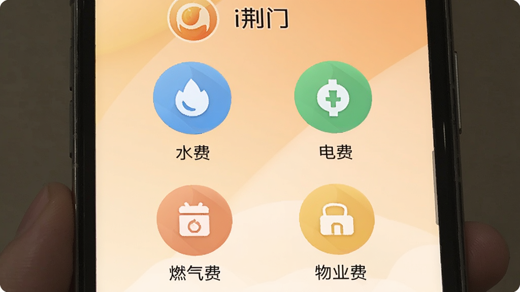 手机屏幕显示“i荆门”生活缴费页面,页面上排列水费、电费、燃气费、物业费四个彩色圆形图标,每个图标下方有文字标签。背景是浅橙色渐变,图标配色柔和,布局为两行两列,整体风格温馨实用,画质干净清晰,无多余元素。