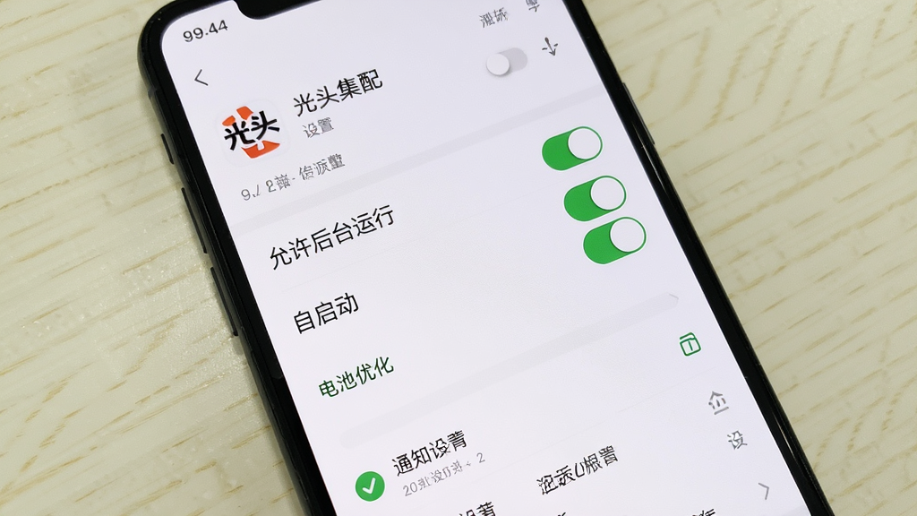 主体为一部手机显示光头集配的设置界面与权限管理页面;布局为手机屏幕特写,背景为浅色桌面;排版上高亮显示“允许后台运行”与“自启动”开关,文字“光头集配”出现在应用名称处;风格为简洁UI截图风格,细节包括权限开关、电池优化选项、通知设置;画质清晰干净,无多余元素。