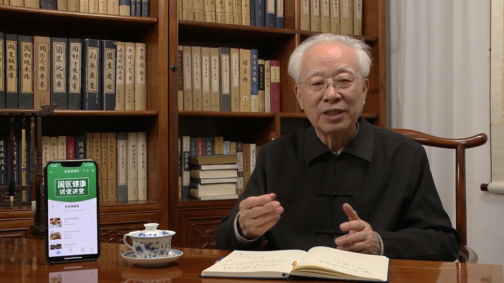 一位白发苍苍的国医大师坐在中式书房中对着镜头讲解养生知识,书架上摆满中医典籍,桌上放着茶杯和笔记本,旁边有一部手机播放“国医健康”应用中的讲堂视频,画面风格温暖亲切,画质清晰干净,无多余元素