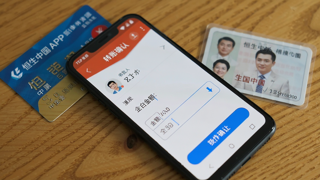 恒生中国APP安卓手机操作界面实拍,手机横放展示转账确认页面,屏幕显示收款人信息与金额输入框,旁边放置一张银行卡与身份证,背景为木质桌面,风格真实自然,画质清晰,干净无多余元素